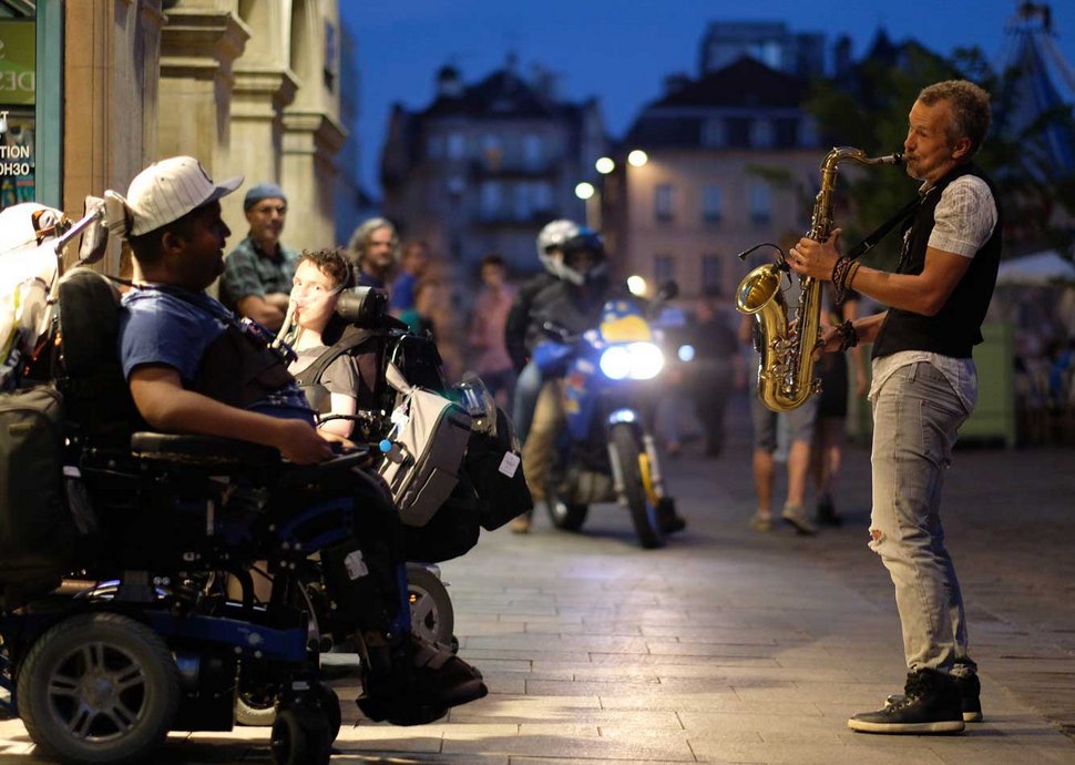 Mann im Rollstuhl hört einem Strassenmusikanten mit Saxophon zu.